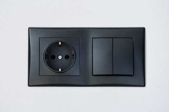 Close up view of a black wallet socket for 220v next to a double light switch against a white wall.