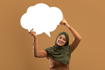 Happy arab lady holding empty speech bubble over her head and smiling at camera over brown...