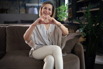 Obraz premium Smiling middle aged woman is showing heart gesture with two hands and looking at camera sitting on sofa.