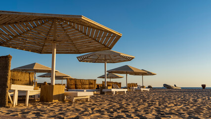 Sunny morning on an Egyptian beach. Lattice umbrellas from the sun against the blue sky. Empty deck chairs are on the sand. An inflatable boat is visible on the shore.