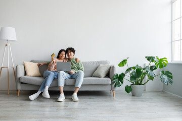 Asian couple using credit card and laptop, sitting on couch, ordering goods in web store, shopping...