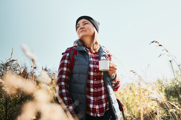 Woman taking break and relaxing with cup of coffee during summer trip. Woman standing on trail and looking away. Woman with backpack hiking through tall grass along path in mountains