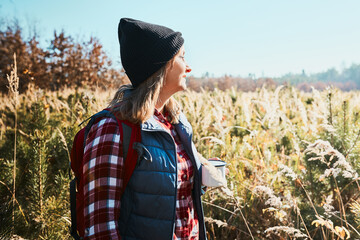 Woman taking break and enjoying the coffee during vacation trip. Woman standing on trail and looking at view. Woman with backpack hiking through tall grass along path in mountains