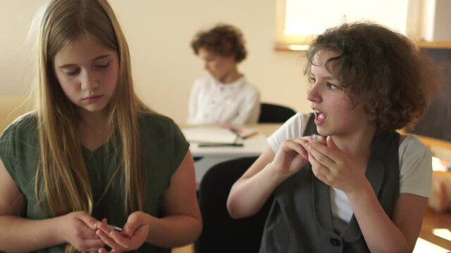 Two busy schoolgirls sit in school class. Their classmate is a curly-haired boy enjoys device free time