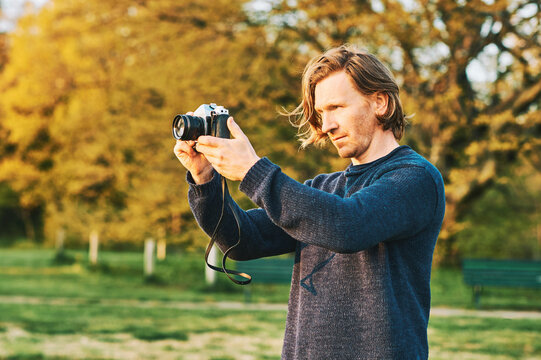 Outdoor portrait of handsome young man taking pictures on green sunny park with old vintage film camera