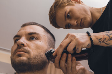 Woman barber uses a hair clipper and styling man's stubble. - obrazy, fototapety, plakaty