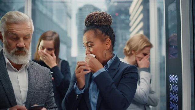 Middle Aged Woman Sneezes in a Crowded Glass Elevator in a Modern Office Building. Businesswoman Covers Her Face, but Other People Are Afraid to Catch the Virus and Microbes in a Lift.