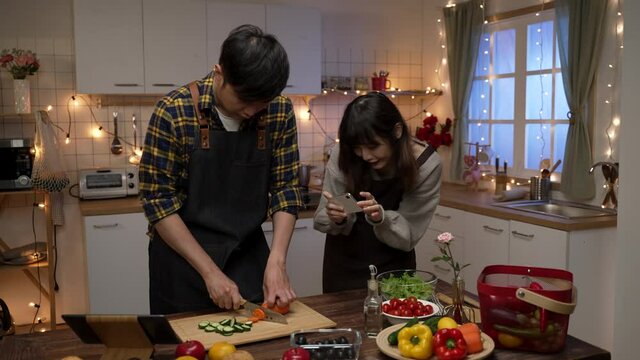 happy asian woman taking picture of her boyfriend making valentine’s day dinner in a romantic kitchen at home. girlfriend shows the man photo she just took on smartphone