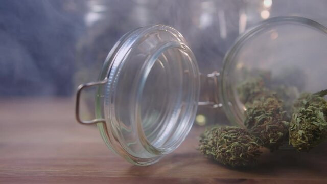 PAN right shot of cannabis buds spilled on the table from a storage glass jar, filled with smoke.