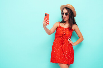 Young beautiful smiling female in trendy summer red dress. Sexy carefree woman posing near blue wall in studio. Positive brunette model having fun in hat and sunglasses. Taking selfie photos