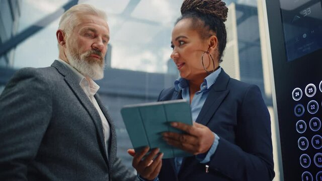 Middle Aged Businessman Talking with International Investment Partner while Riding Glass Elevator to Office in a Modern Business Center. Corporate Associates Use Tablet Computer in a Lift.