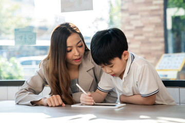 Businesswoman sitting beside kid and teach homework in the coffee shop or cafe on summer. Boy looking at tablet and smile. Feeling happy during learning. Education and weekend concept.