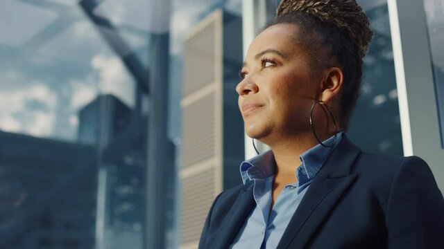 Portrait of Successful Black Businesswoman Riding Glass Elevator to Office in Modern Business Center. African American Female Looking at Modern Skyscrapers Out of the Panorama Window in the Lift.