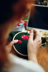 Redhead woman embroidered mushroom hat in hoop. on green material. Wooden table with laptop on background. Concept of needlework, hobby, leisure. Close-up.