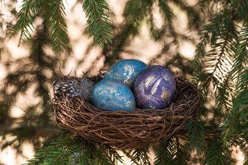 Happy Easter 2022. A postcard with a copy of the place for the text. Purple, blue and golden eggs in a nest on a natural forest background. Top view.