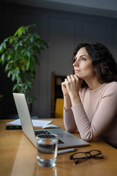 Young thoughtful female employee sitting at office desk with laptop looking through window, thinking about future or dreaming of vacation. Pensive dreamy businesswoman with hands under chin at work