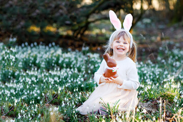 Adorable little girl with Easter bunny ears eating chocholate figure in spring forest on sunny day,...