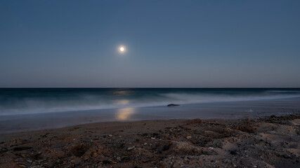 A night on an Egyptian beach. The full moon is shining in the dark blue sky. Waves are foaming on the sand. Moonlight path on the water. Long exposure