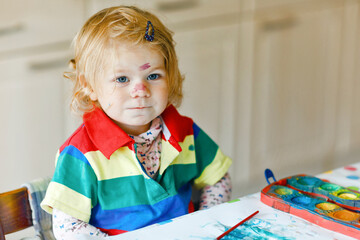 Cute adorable baby girl learning painting with water colors. Little toddler child drawing at home, using colorful brushes. Healthy happy daughter experimenting with colors, water at home or nursery.