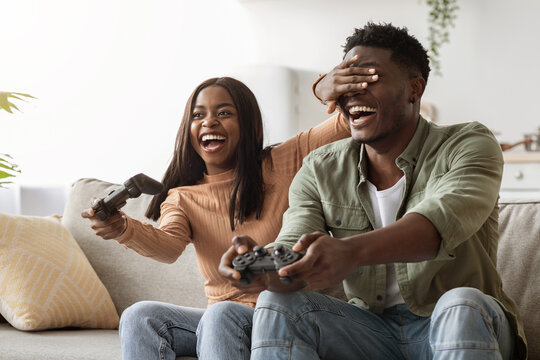 Laughing black woman closing boyfriend eyes, playing video games