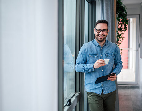 Portrait of young adult smiling cheerful businessman in home office drinking coffee morning routine starting day happy handsome successful entrepreneur freelancer checking his business tasks for day