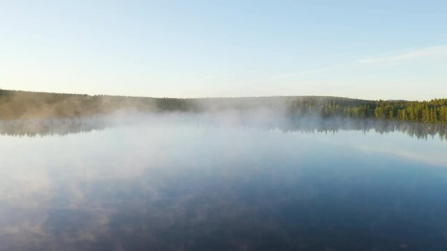Flying over a blue foggy lake during sunrise, camera slowly turning towards the sun with green forest in the background filmed in Lapland Finland.