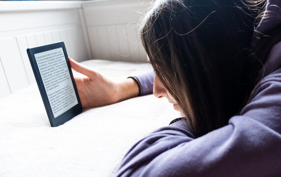 Young Adolescent GIrl is focused reading an electronic romance with her Ebook Reader while she is on the bed. The text on the ebook reader is a "lorem ipsum"