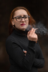 A beautiful girl in a dark blouse smokes a cigarette outdoors.