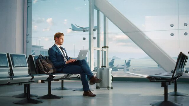 Modern Airport Terminal: Handsome Businessman Working on Laptop Computer While Waiting for His Flight. Man Sitting in a Boarding Lounge of Big Airline Hub with Airplanes Departing and Arriving