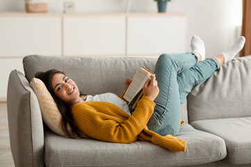 Carefree middle eastern woman resting on couch in living room, reading book while resting at home,...