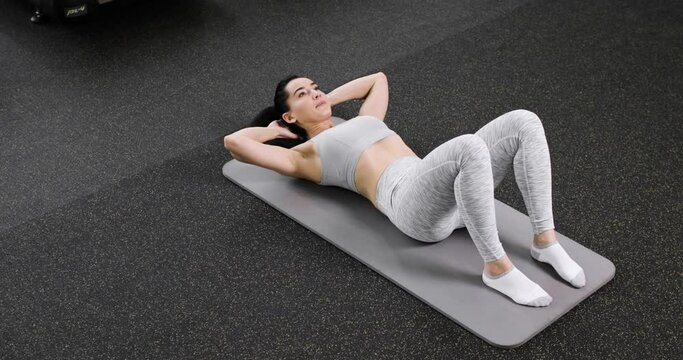 A young athletic woman does an exercise on the abdominal press, lies on the floor in the gym, dressed in gray sportswear. Top view. Active lifestyle concept.