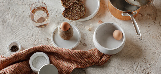 Obraz na płótnie Canvas Breakfast setting with boiled egg in stoneware egg cup, whole grain rye bread, glass of water, salt flakes and pepper in concrete bowls on rough textured clay background. Healthy breakfast concept