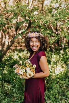Smiling woman stands in woods with bouquet and wearing floral crown