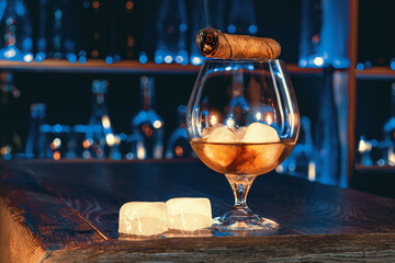 Cigar, elegant glass of brandy on the bar counter. Alcoholic drinks, cognac, whiskey, port, brandy, rum, scotch, bourbon. Vintage wooden table in a pub at night