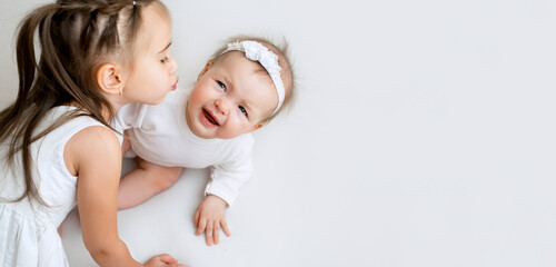 Six-month-old baby sobs avoiding the kisses of his little sister. Relationship of children in a family with an age difference. Girls in white clothes on a white studio background with a copy space
