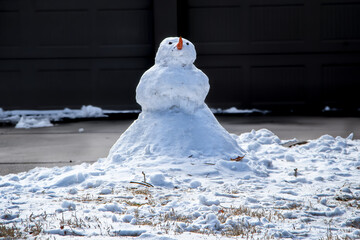 Snowman with crossed arms and carrot nose in front of dark wall and drive with snow in foreground.