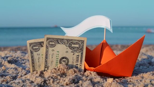 Two paper one dollars bills half buried in sand, orange paper boat with and empty blank speech bubble for text close-up on sandy beach near sea. Concept money rest vacation traveling tourism finance