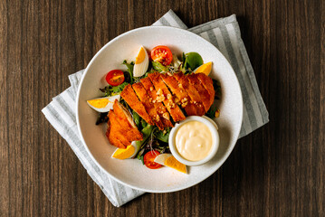 Chicken schnitzel served with fresh salad, boiled egg, tomato and mayo in white bowl on dark wood background top view.