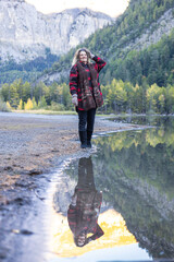 Full body portrait of happy smilling woman posing at lake. Nature mountain swiss alps background. Natural lighting and water reflection. Warm autumn clothes.