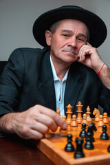 Portrait of smilling man playing chess, holding his head. Classical formal suit and hat, vintage look. Happy chess player making move. Competitive strategy leisure game.