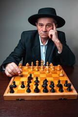 Portrait of man with hat sitting, moving chess piece. Classical formal suit, vintage look. Chess player posing with board, holding his head thinking.