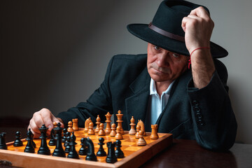 Portrait of man sitting with chess board, tipping his hat to the camera. Classical formal suit, vintage look. Dark background with dim light.
