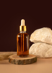 Transparent glass cosmetic dropper standing on wooden podium near stones and dry flowers on dark beige background. Beauty composition. Concept of natural and original skincare products presentation. 