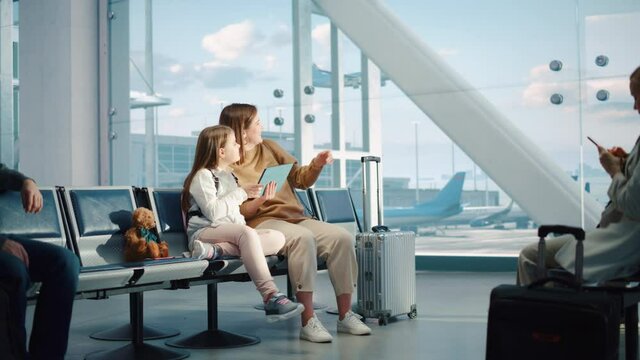Busy Airport Terminal: Cute Mother and Little Daughter Wait for their Vacation Flight, Play Educational Games on Digital Tablet. People Sitting in Boarding Lounge of Airline Hub with Airplanes Flying