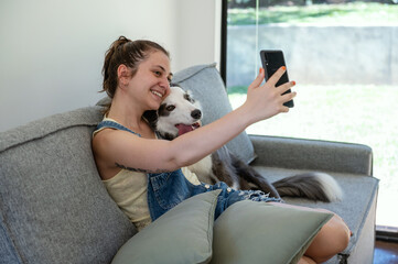 young woman hugging her border collie dog on the sofa and taking selfie