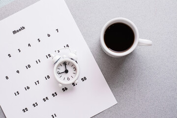 Blank sheet of monthly calendar, coffee cup and alarm clock  on a gray background. Time planning and organization. Copy space. Close-up