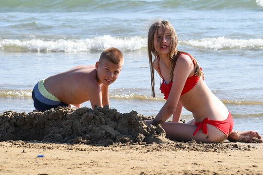 children play on sand by sea. teenagers are sitting on seashore in swimsuits. kids fun holidays in tropics in summer