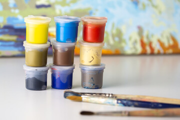 Small jars filled with paint on a painted canvas background. Multi-colored cans of paint, brushes lie nearby. Hobby concept, drawing. Painting by numbers.
