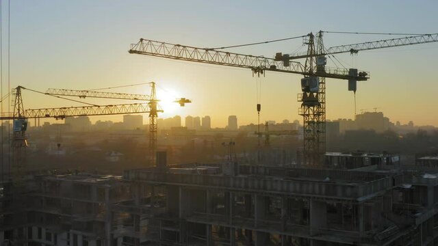 Aerial view of a large construction of a residential area with many multi-storey buildings under construction, a lot of tower cranes, workers, construction equipment at sunset