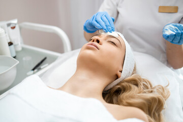 Female cosmetologist in medical gloves, applying cream or foam on a woman's face to clean her skin. Beautician doctor making anti-age procedure